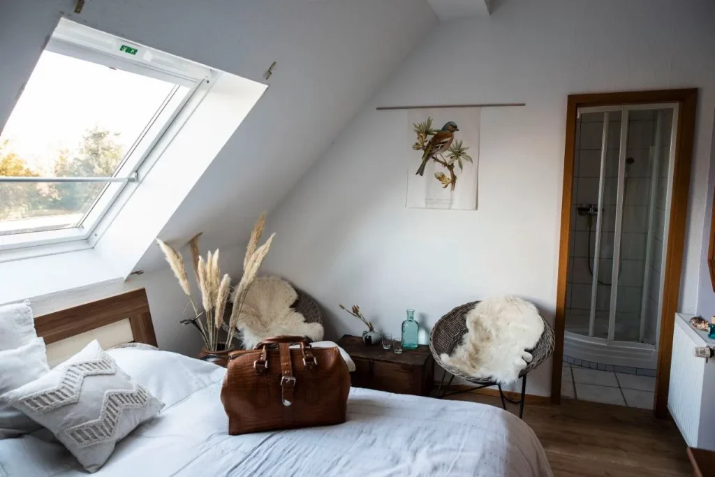 Gemütliches Schlafzimmer mit Bett, zwei Stühlen, Pflanzen und einem braunen Sitzkoffer.