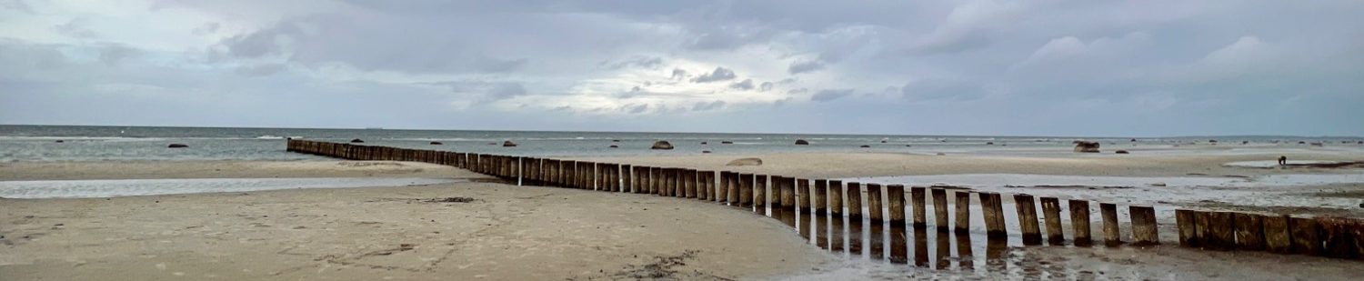 Sandstrand mit Steg und Wolken am Himmel, weites Meer im Hintergrund.