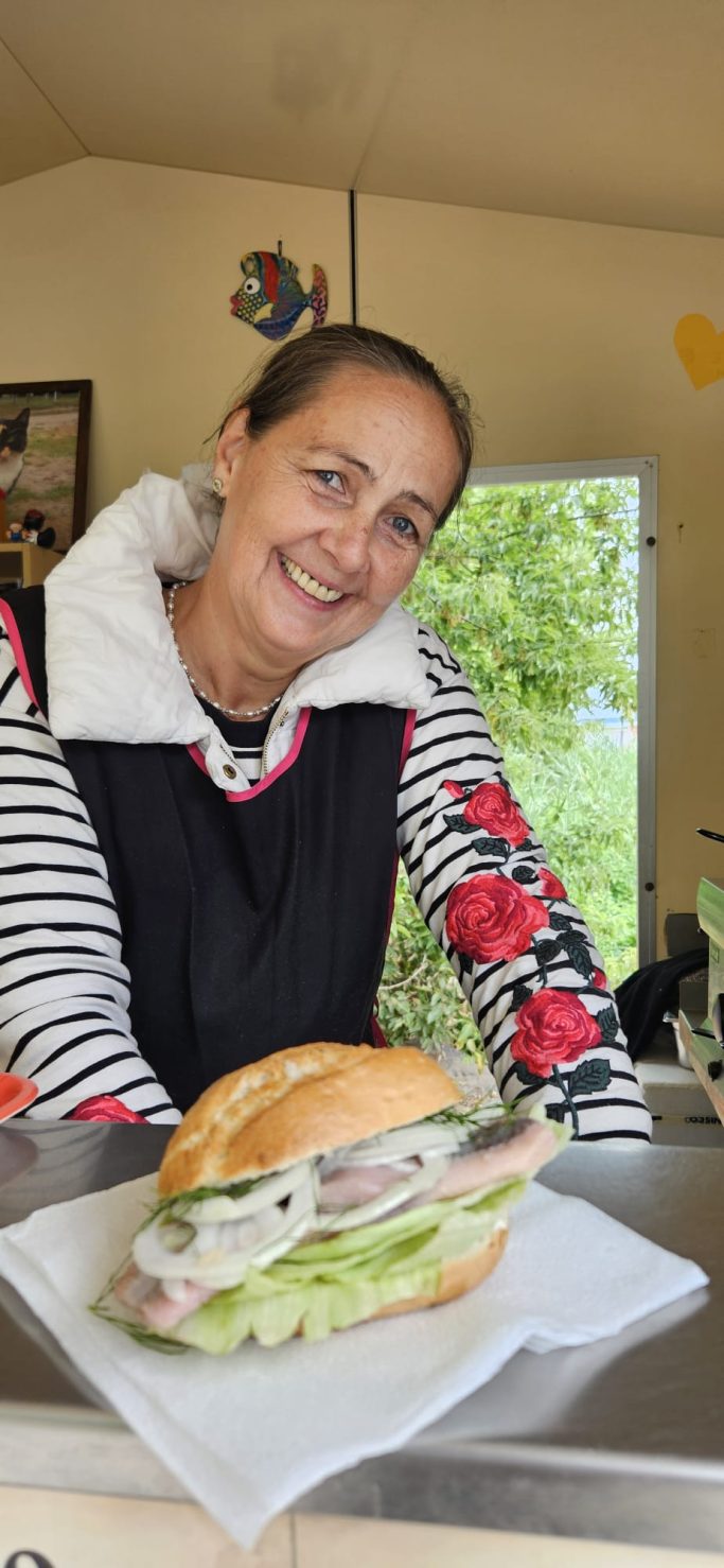 Lächelnde Frau mit Sandwich auf einem Tisch, umgeben von natürlichem Licht.