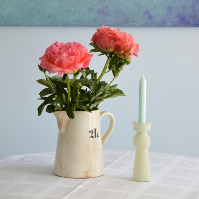 Rosa Pfingstrosen in einer weißen Kanne neben einer grünen Kerze.