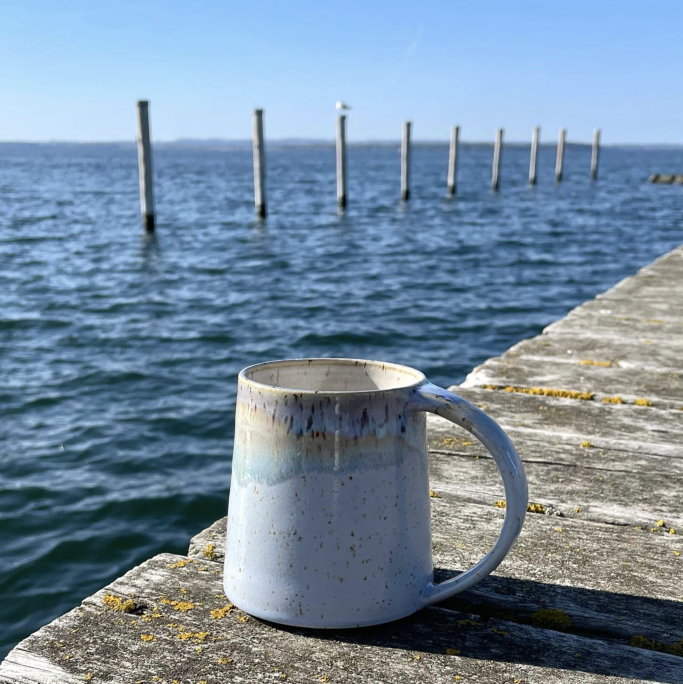 Eine Tasse steht auf einem Holzsteg am Wasser, im Hintergrund Pfahlreihen.
