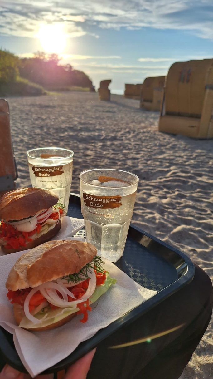 Zwei Gläser Wasser und Sandwiches auf einem Tablett bei Sonnenuntergang am Strand.