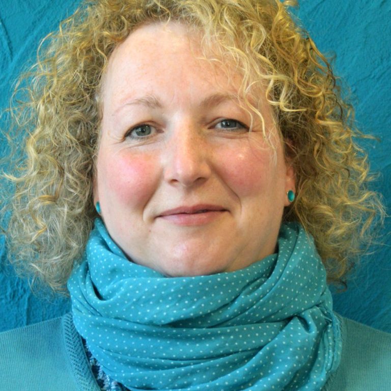 Frau mit lockigem, blondem Haar und blauem Schal vor einer türkisfarbenen Wand.