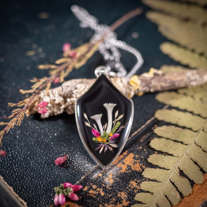 Schwarzes Anhänger mit floralem Design auf silberner Kette, umgeben von Naturelementen.