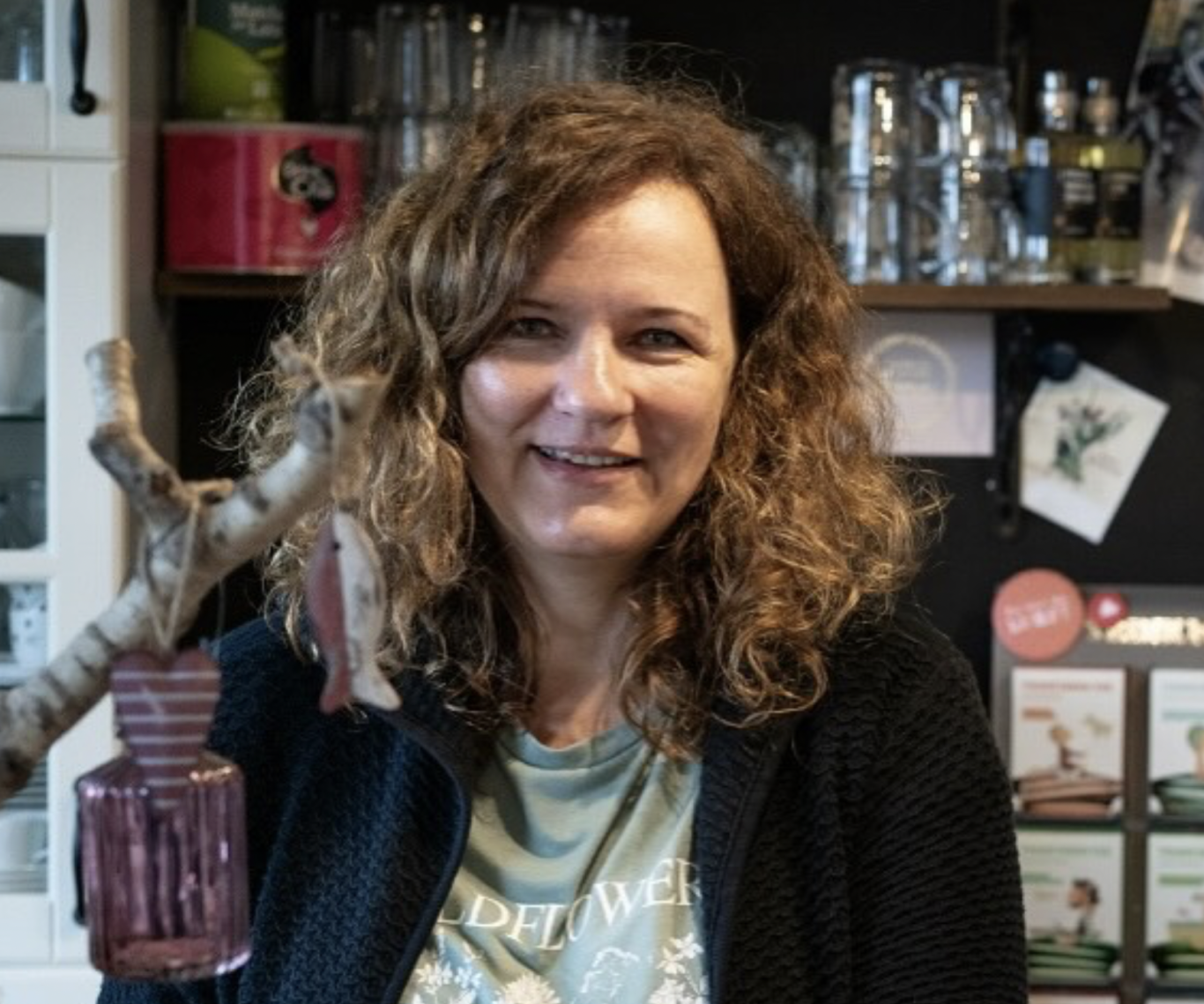 Frau mit lockigem Haar lächelt in einer gemütlichen Umgebung mit Glasobjekten.