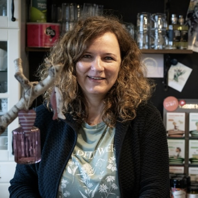 Frau mit lockigem Haar lächelt hinter einer Theke, umgeben von Flaschen und Dekoration.