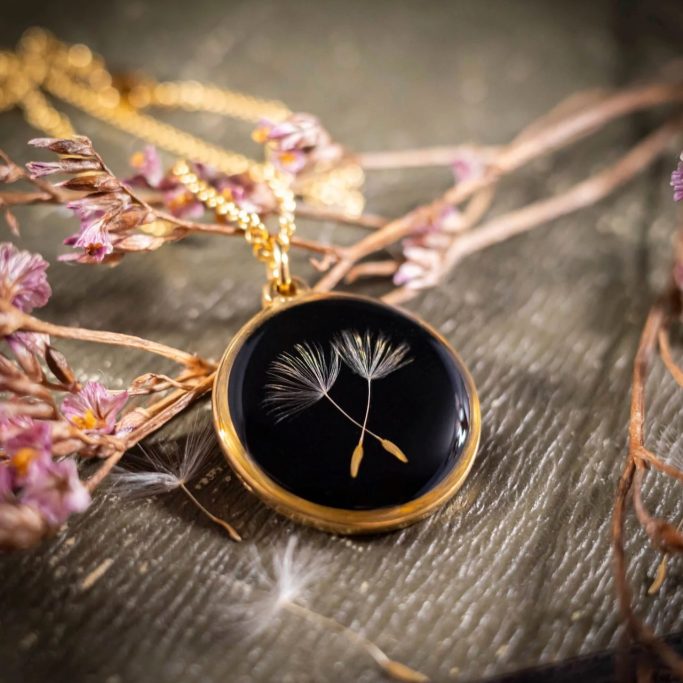 Schwarze Halskette mit goldener Fassung, die zwei Dandelion-Samen zeigt.