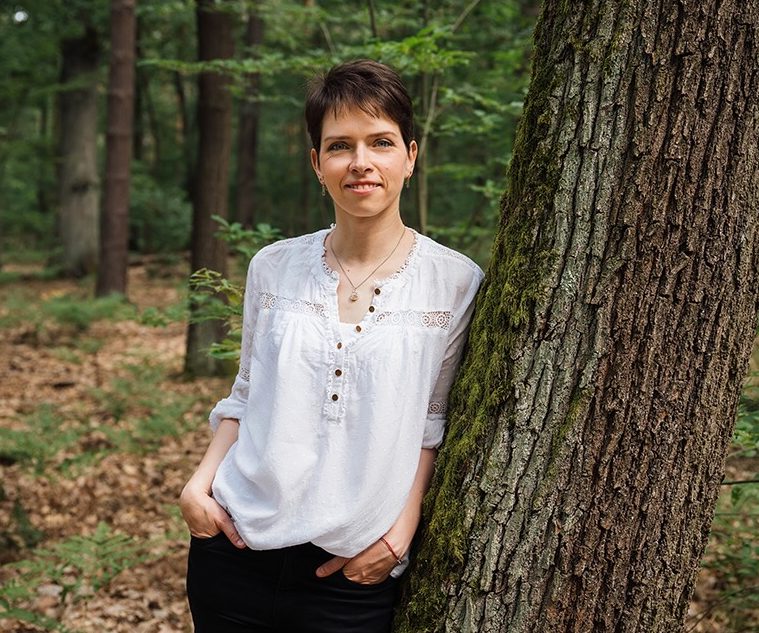 Frau steht selbstbewusst neben einem Baum im Wald, trägt ein helles Oberteil.