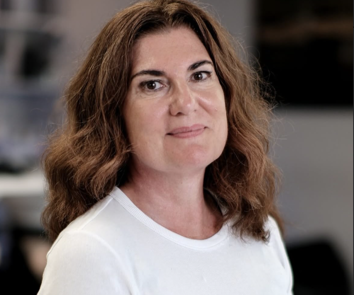 Frau mit mittellangen braunen Locken, lächelt freundlich in lässiger Kleidung.