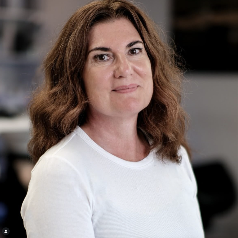 Frau mit mittellangen, lockigen Haaren, trägt ein weißes Shirt und schaut freundlich.