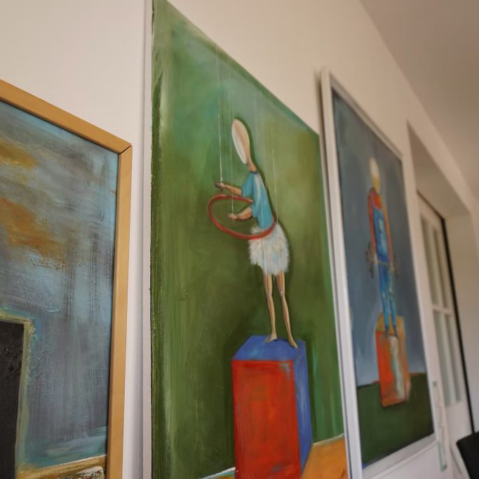 Drei farbenfrohe Gemälde an einer Wand, zwei Figuren mit Reifen und Kästen.