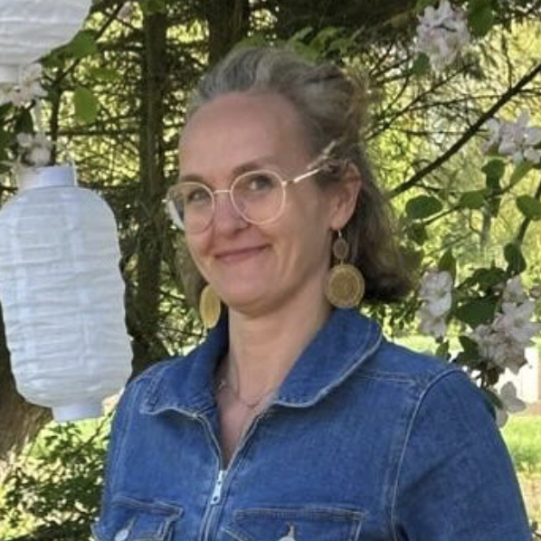 Frau mit Brille und Ohrringen, trägt eine Jeansjacke, steht vor einem Baum mit Laternen.