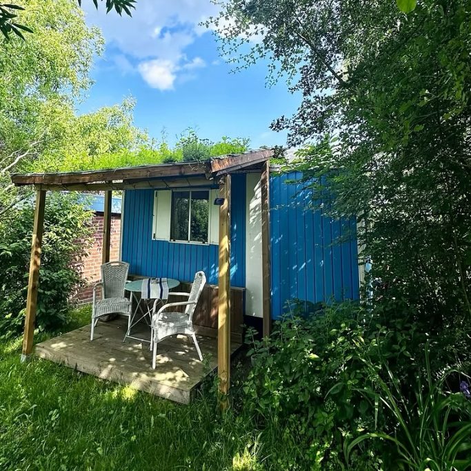 Kleine blaue Hütte umgeben von Grün, mit einem überdachten Sitzbereich und hellblauem Himmel.