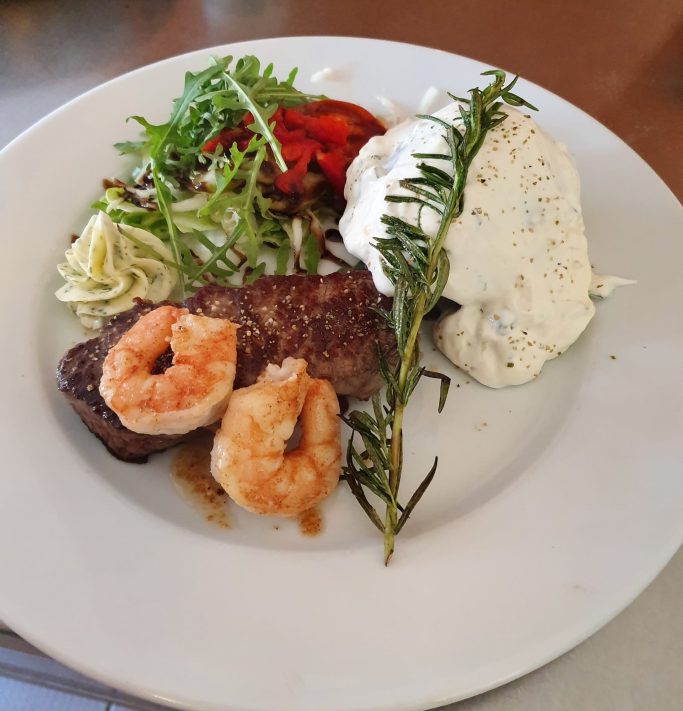 Ein Teller mit Steak, Garnelen, Salat und einer weißen Sauce, garniert mit Rosmarin.