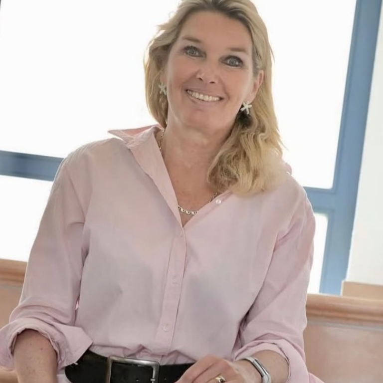 Frau mit blonder Haarfarbe in einem rosa Hemd lächelt am Fenster sitzend.