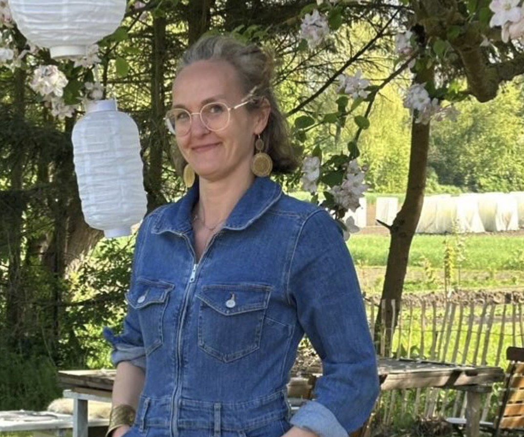 Frau in Jeansjacke steht im Freien, umgeben von Blumen und Laternen.