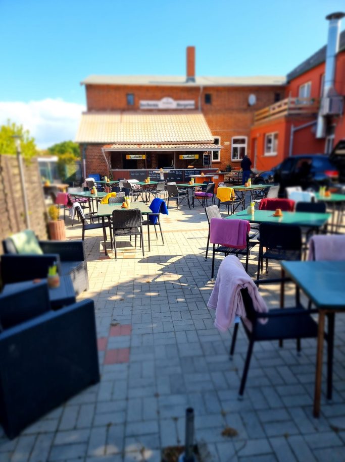 Gemütlicher Außenbereich eines Cafés mit Tischen, Stühlen und bunten Blumendekorationen.