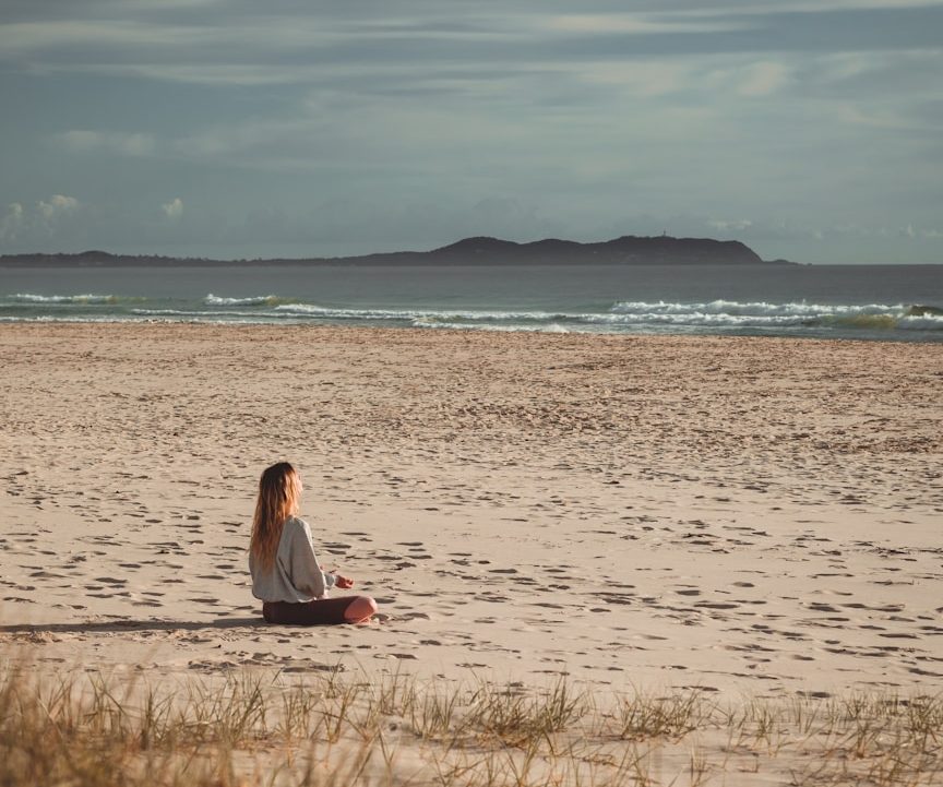 Frau sitzt alleine am Strand, Blick auf das Meer und eine Insel im Hintergrund.