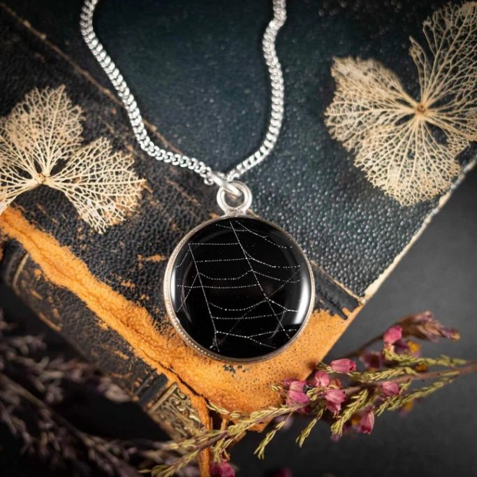 Eine silberne Halskette mit einem runden Anhänger und schwarzem Stein und Spinnennetz-Muster.