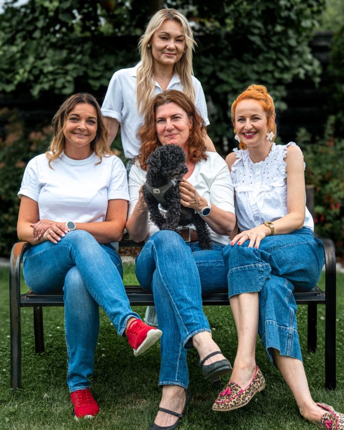 Vier Frauen mit einem Hund sitzen auf einer Bank im Freien, umgeben von grüner Landschaft.