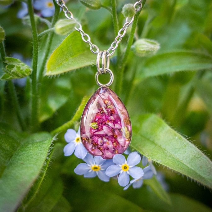 Tropfenförmiger Anhänger mit getrockneten Blumen, umgeben von Vergissmeinnicht-Blüten.