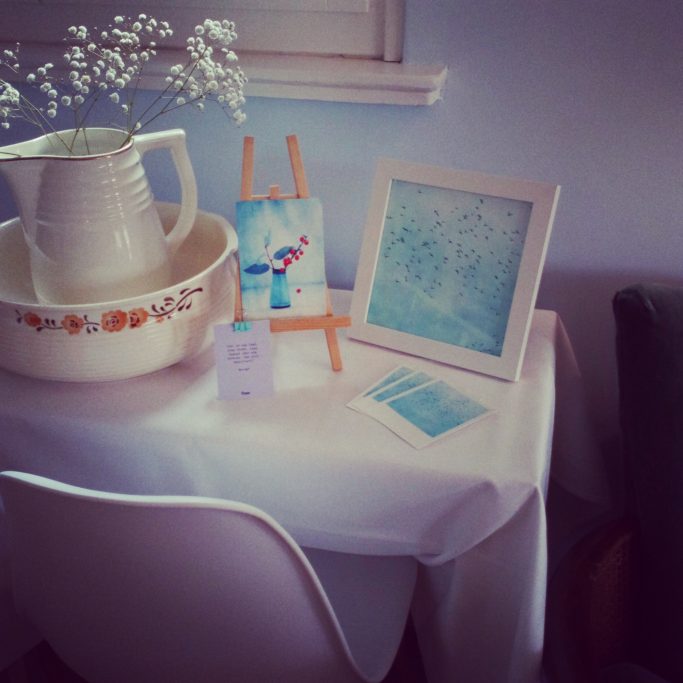 Ein Tisch mit einer weißen Kanne, einem Gemälde und einem gerahmten Bild in sanften Blautönen.