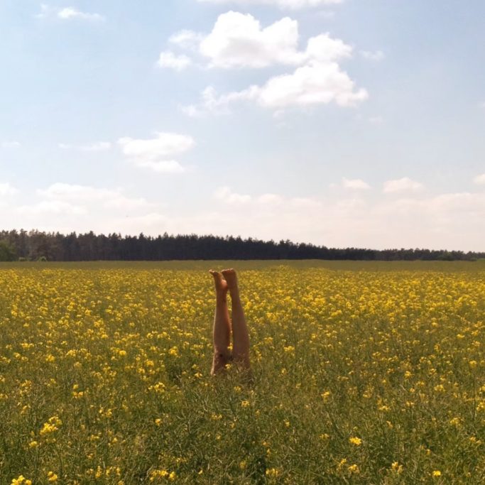 Eine Person mit Beinen nach oben in einem blühenden, gelben Feld unter blauem Himmel.