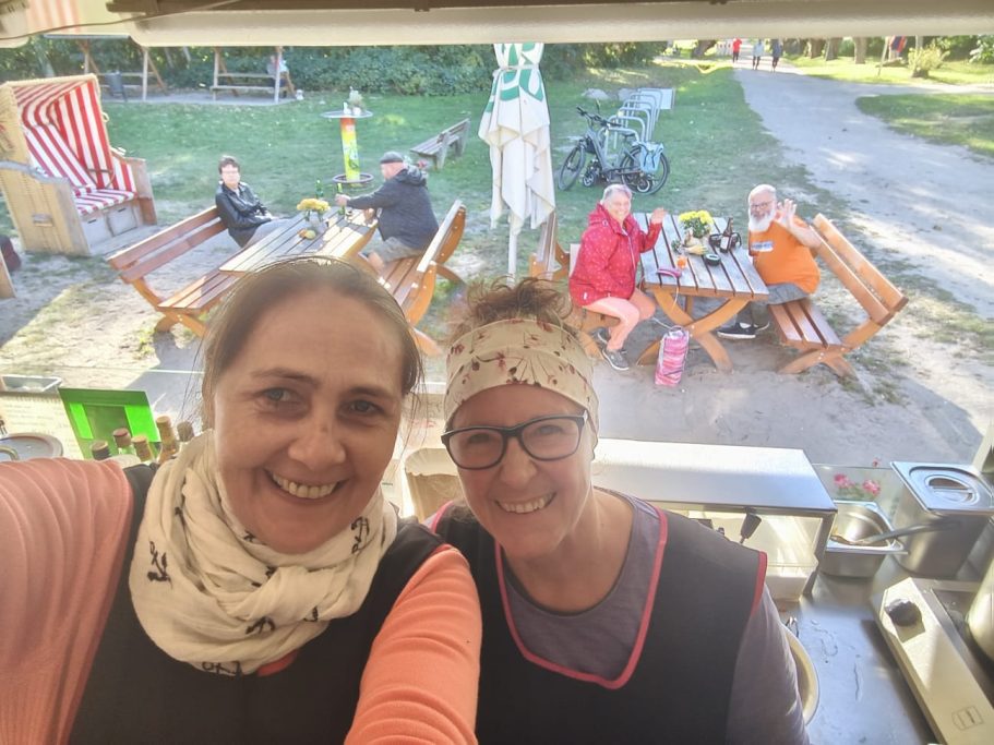 Zwei Frauen lächeln in einem Freiluftcafé, im Hintergrund sitzen andere Gäste.