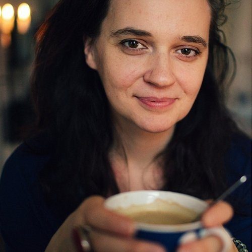 Lächelnde Frau mit langen Haaren, schwarzem Oberteil und blauer Strickjacke.