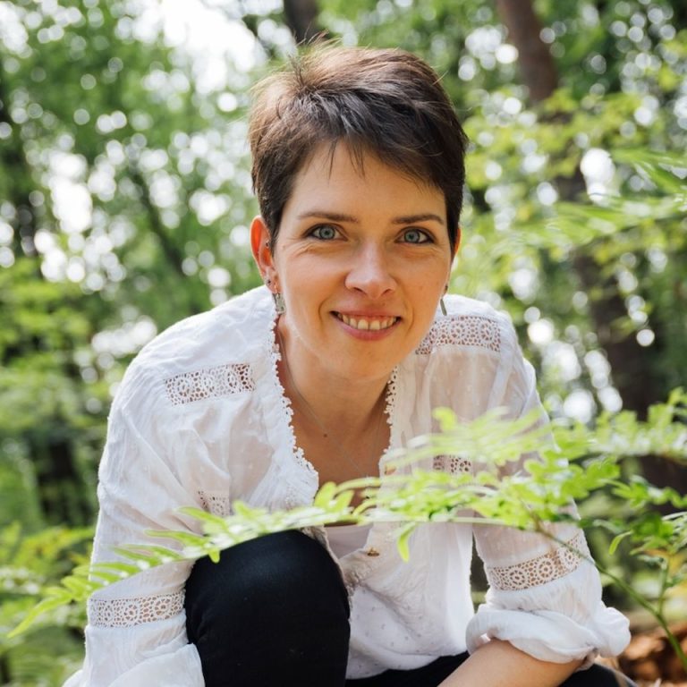 Frau in einem weißen Oberteil kniet zwischen Pflanzen im Wald und lächelt.