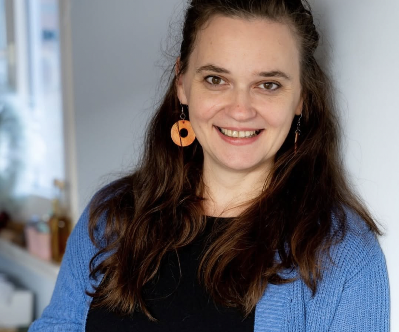 Lächelnde Frau mit langen Haaren, orangen Ohrringen und blauer Strickjacke.