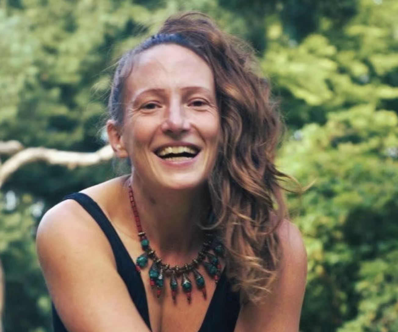 Junge Frau mit lockigem Haar und Halskette lächelt vor grünem Hintergrund.