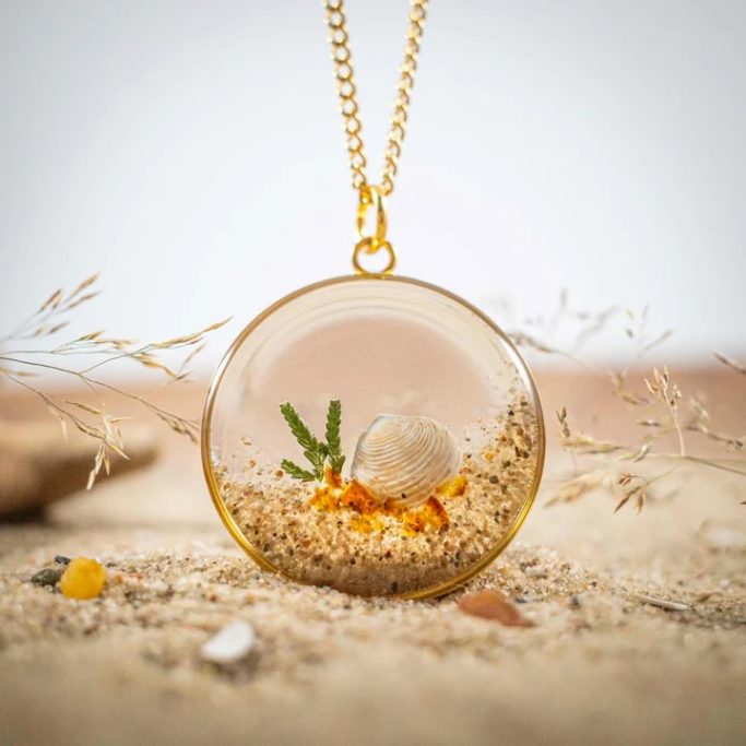 Ein goldener Anhänger mit einer Sandlandschaft und kleinen Pflanzen im Inneren.