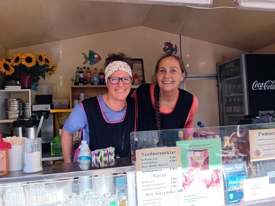 Zwei Frauen lächeln hinter einem Thekenstand mit Getränken und Informationsflyern.